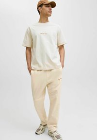 Creme bomulds T-shirt og matchende joggers med diskret broderi, båret med en tan kasket og beige sneakers. Enkelt, afslappet fits design.