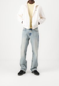 White lightweight jacket, yellow t-shirt, light blue jeans, black sneakers. Jacket has a zip closure and elastic cuffs. Jeans have a relaxed fit.