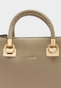 Sac à main en cuir beige avec une poignée tressée, des éléments en métal doré et un logo détaillé. Texture lisse avec un design structuré.
