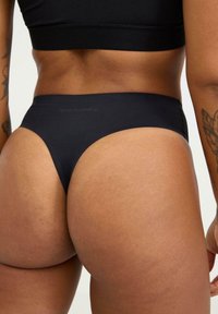 Schwarzer Tanga aus einem glatten, dehnbaren Stoff mit hohem Beinausschnitt, minimalistischem Branding und nahtlosen Kanten.