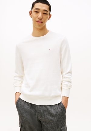 Jeune homme portant une chemise blanche à manches longues avec un petit logo et un pantalon gris texturé, debout les mains dans les poches sur un fond neutre.