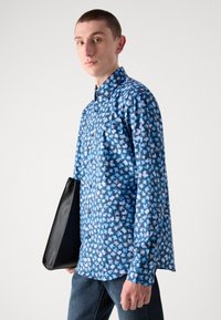 Chemise à boutons à motif floral bleu avec des manches longues, en tissu léger. Porte-documents en cuir noir tenu dans la main gauche.