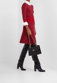 Femme portant une robe rouge, des poignets de chemise blancs, des bottes noires à talons hauts jusqu'au genou, et portant un sac à main noir avec des poignées en chaîne dorée.