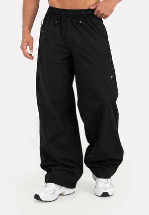 Hombre con pantalones cargo negros holgados con cintura elástica y zapatillas blancas, de pie frente a un fondo liso.