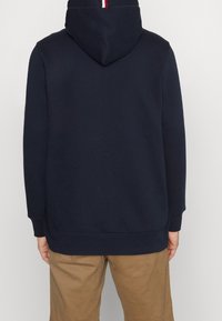 Donkerblauwe hoodie met capuchon en trekkoord, geribde manchetten en zoom. Heeft een verticale rood-witte streep achter in de nek. Zachte textuur.