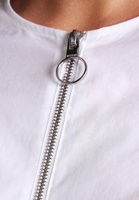 Indumento bianco con chiusura a zip argentata dotata di un tirante circolare, evidenziato da nastro della zip testurizzato e trama di tessuto liscia.