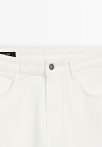 Pantaloni in denim bianco con chiusura a bottone, superficie testurizzata e dettagli di cucitura strutturati lungo la vita e le tasche.