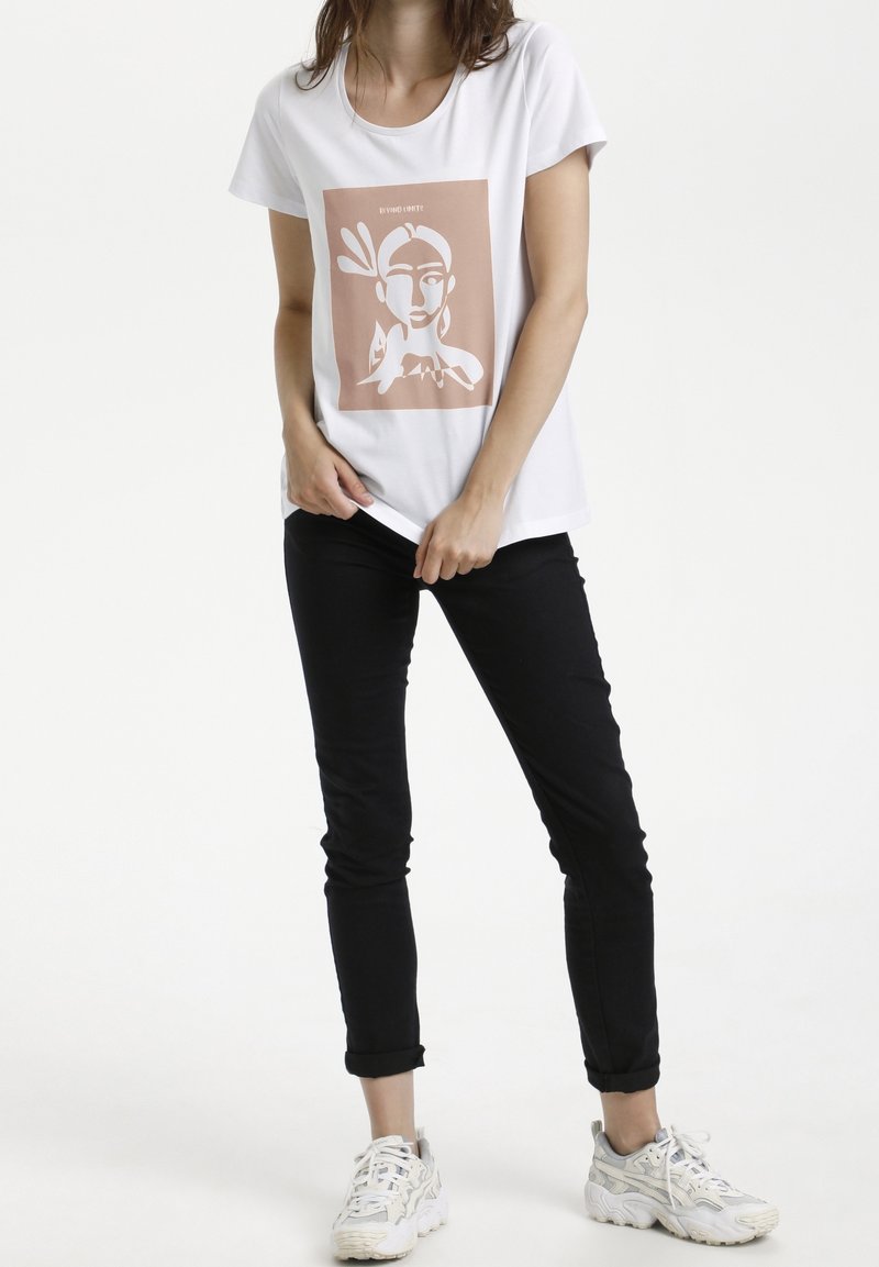 Femme portant un t-shirt blanc à motif, un pantalon noir retroussé et des baskets blanches, debout devant un fond blanc.