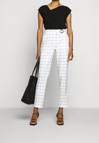 Haut noir à manches courtes avec un détail drapé, pantalon blanc taille haute avec un motif à carreaux noir, sac cabas noir, et sandales à brides.