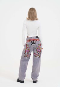 Frau mit schulterlangem blonden Haar trägt ein weißes Langarmoberteil und graue Jeans, die auf der Rückseite mit buntem Drachen- und Dolchstickereien verziert sind.