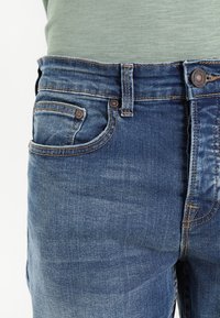 Gros plan de la taille d'un jean en denim bleu avec poches avant, passants de ceinture, bouton, et chemise verte rentrée.