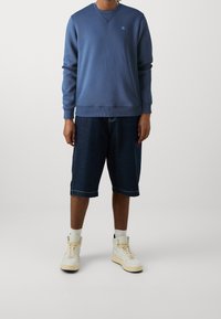 Felpa blu con scollo rotondo, abbinata a pantaloni corti in denim fino al ginocchio. Indossata con sneakers bianche da atletica e calzini alla caviglia.