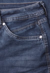 Primer plano de unos vaqueros de mezclilla azul oscuro que muestran el bolsillo delantero, el pequeño bolsillo para monedas, detalles de costura y remaches metálicos.