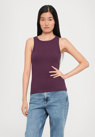 Aangemeten, mouwloos, paarse tanktop met geribbelde structuur en een ronde halslijn. Bevat een subtiel logo op de borst. Gepaard met lichtblauwe jeans.