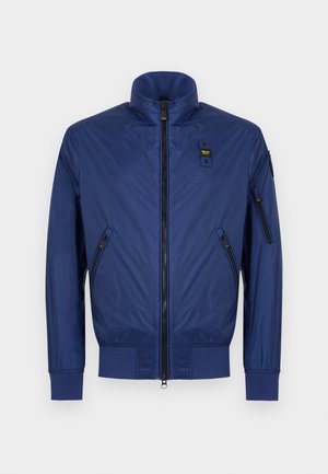 Granatowa kurtka na zamek z kołnierzem typu stójka, dwoma przednimi kieszeniami na zamek, mankietami na nadgarstkach oraz małą naszywką z logo na piersi i kieszeni na rękawie.