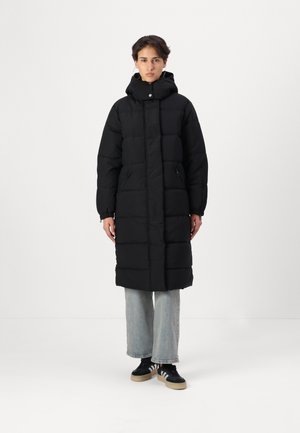 Zwarte pufferjas met een capuchon, voorzien van een doorgestikt ontwerp, een rits aan de voorkant, twee zijzakken en elastische manchetten. Gedragen over lichtblauwe jeans.