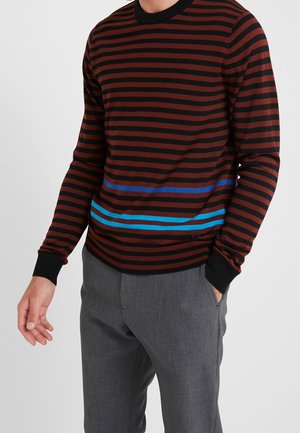 Mężczyzna w czarnym i ciemnoczerwonym swetrze w paski z niebieskimi paskami w okolicy talii, ubrany w szare spodnie, stoi z jedną ręką w kieszeni.