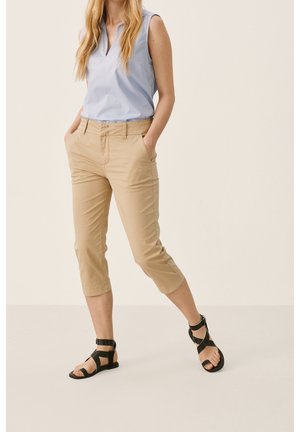 Femme vêtue d'un chemisier sans manches bleu clair, de pantalons beige courts et de sandales plates noires à lanières, debout les mains dans les poches.