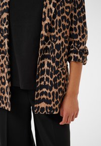 Blazer con stampa leopardata in tessuto oro e nero, caratterizzato da maniche arrotolate e una tasca frontale singola, abbinato a un top e pantaloni neri.