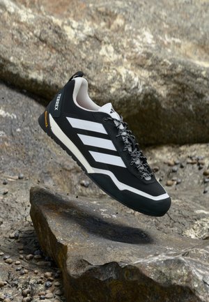 Black athletic shoe with white stripes, textured upper, and sturdy rubber sole. Features reinforced toe and intricate lace design.