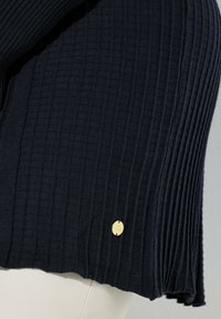 Nærbillede af en sort ribstrikket nederdel med en lille rund guldtags nær kanten, båret over beige bukser.