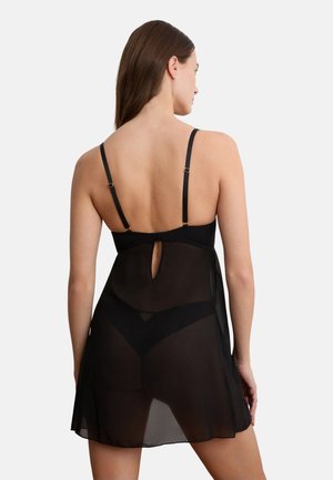 Femme aux cheveux bruns portant une robe de lingerie noire transparente et des sous-vêtements assortis, vue de dos sur un fond clair uni.