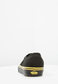 Musta kangaskengät matalalla profiililla, keltainen raita pohjassa ja kumipohja. Ominaisuuksiin kuuluu pyöreä kantapää ja logo takana.