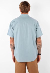 Chemise bleu clair à manches courtes avec un tissu texturé, un col et une étiquette dans le dos. Portée avec un short beige, révélant des bras tatoués.