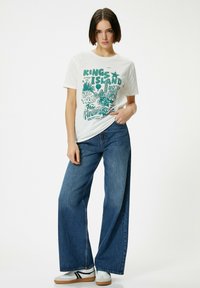 Hvid grafisk t-shirt med grøn print, parret med vidde jeans i blå denim. Hvide sneakers med sorte accenter fuldender outfittet.
