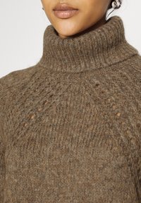 Gros plan d'une personne portant un pull à col roulé marron tricoté avec des motifs texturés et des boucles d'oreilles en argent en forme d'anneau.