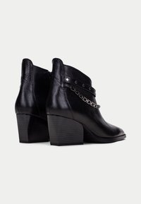 Botas de cuero negro hasta el tobillo con un acabado texturizado, que presentan una cadena decorativa y una correa con tachuelas, además de un tacón sólido y cuadrado.