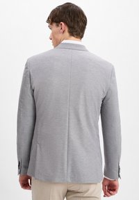 Helles graues Blazer aus strukturiertem Stoff, schlanke Passform, taillierte Revers und zwei Rückenbelüftungen; kombiniert mit beigen Hosen.