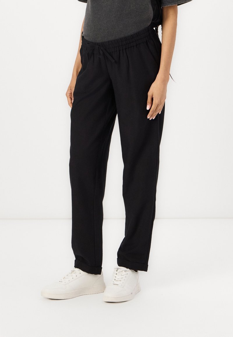 Personne portant un pantalon noir à cordon et des baskets blanches, debout devant un fond blanc uni.