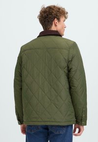 Giacca trapuntata verde oliva con colletto in cotone corduroy marrone. Presenta un motivo a diamante, dettagli con bottoni sulle maniche e una vestibilità rilassata.