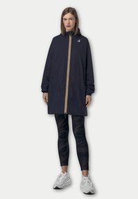 Veste coupe-vent marine avec des rayures jaunes et blanches, fermeture zippée, capuche et poches latérales, associée à des leggings noirs et des baskets blanches.