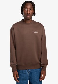 Sudadera marrón con cuello redondo y puños de canalé. Presenta un logotipo en texto blanco en el pecho. Combinada con jeans de mezclilla azul.