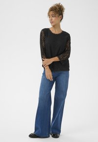 Sort blondeløsts top med 3/4 ærmer, rund halsudskæring, lagdelt over lyseblå, vide jeans, parret med sort fodtøj. Glat tekstur.