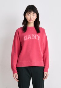 Czerwona bluza z logo "GANT" w jasnoróżowym kolorze, z okrągłym dekoltem, ściągaczami na rękawach i u dołu, noszona na ciemnozielonych spodniach.