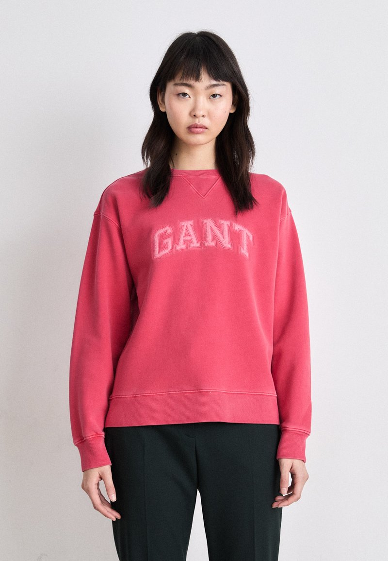Czerwona bluza z logo "GANT" w jasnoróżowym kolorze, z okrągłym dekoltem, ściągaczami na rękawach i u dołu, noszona na ciemnozielonych spodniach.