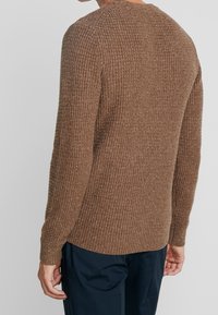 Man iförd en brun strukturerad stickad tröja och mörkblå byxor, visad från ryggen mot en enhetlig bakgrund.