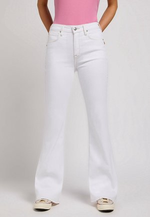 Kvinde iført hvide højtaljede flare jeans, lyserød ærmeløs top og cremefarvede casual sneakers, med hænderne hvilende bag ryggen.