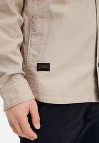 Beige Jacke aus glattem Stoff, mit einem schwarzen Etikett und goldener Schrift. Nahaufnahme des Ärmeldetails zeigt Naht und Manschetten.
