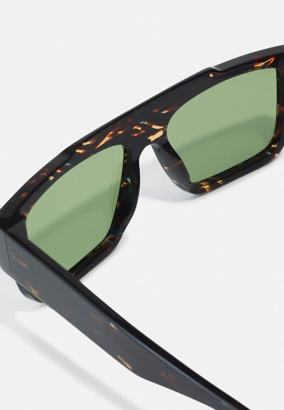 Gafas de sol con montura negra y carey, lentes verdes y diseño angular. Presentan un acabado brillante y acentos prominentes en las patillas.