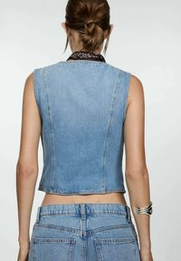 Veste courte en jean sans manches, de couleur bleu clair, avec des détails de coutures apparentes. Associée à un bas en jean bleu clair.