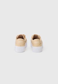 Beige Lacoste kedas, skatītas no aizmugures, ar baltām biezām zolēm un "LACOSTE" zīmolu uz papēža.