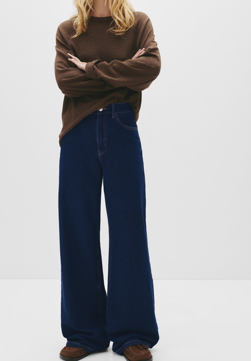 Chemise marron à manches longues associée à un jean bleu foncé à jambe large. Le denim présente des coutures visibles et un design classique à cinq poches. Chaussures brunes incluses.