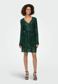 Zelena obleka s tkanimi bleščicami, dolgimi rokavi, V-izrezom in zategnjenim pasom. Ima krilo s širokim robom in je v paru s črnimi čevlji na visokih petah.