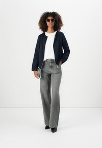 Marineblau gefütterte Bomberjacke, weißes T-Shirt, graue weit geschnittene Jeans, schwarze Knöchelstiefel und Sonnenbrille im Cat-Eye-Stil. Neutraler Hintergrund.