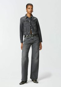 Tamni traper jakna skraćena do struka s kopčanjem na dugmad i dva dzepa na grudima, u kombinaciji s širokim tamnim traper hlačama i pojasom sa srebrnom kopčom.