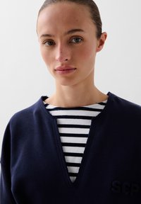 Marineblauwe sweatshirt met v-hals, gemaakt van een gebreide stof met rauwe randen, gedragen over een zwart-wit gestreept shirt. "SCP" geborduurd.